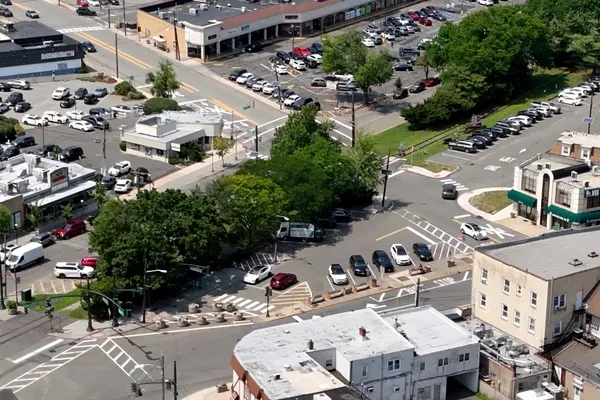 Aerial view of a city street with cars parked on the side of it, surrounded by buildings, trees, poles, lights